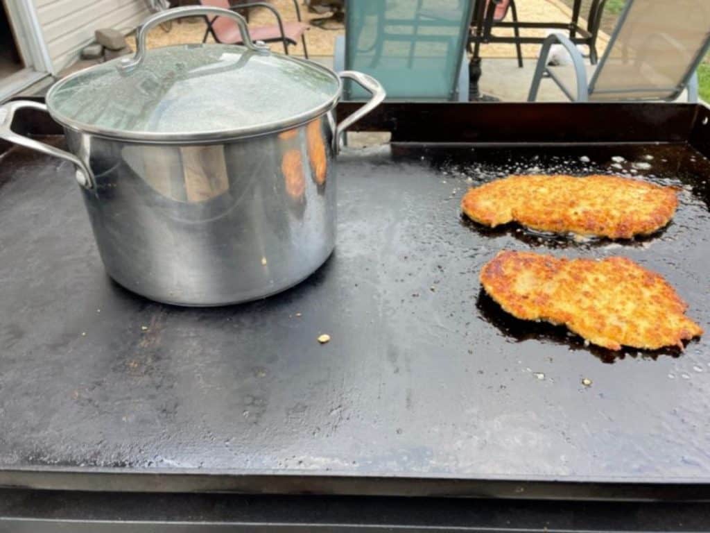 A pot of water boiling on the blackstone griddle while chicken cutlet cook next to it