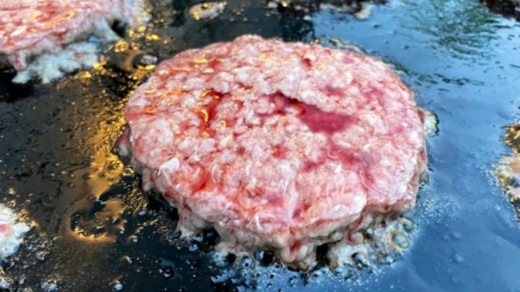 A frozen burger patty cooking on a blackstone griddle and ready to be flipped