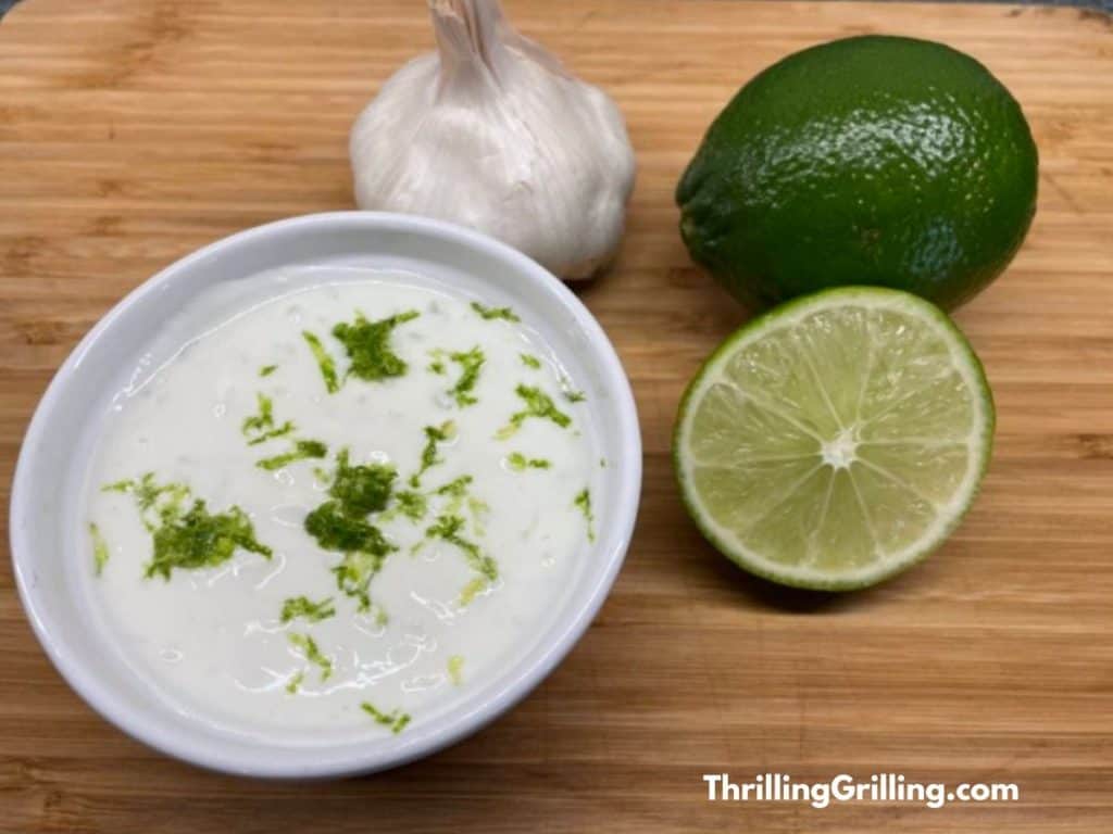 A bowl of garlic lime crema with some fresh limes and a bulb of garlic.