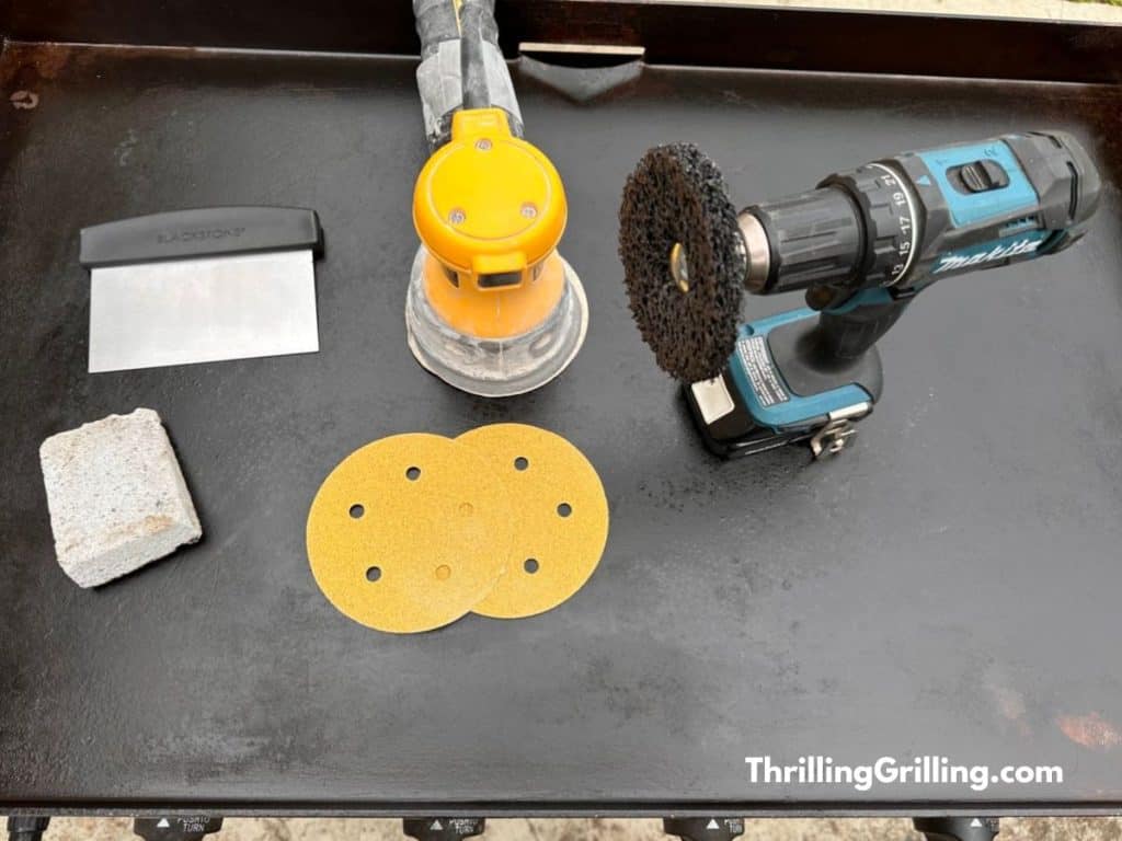 All the tools you need to strip and re-season a blackstone griddle including a grill brick, an orbital sander, and a power drill with a scrapper bit.