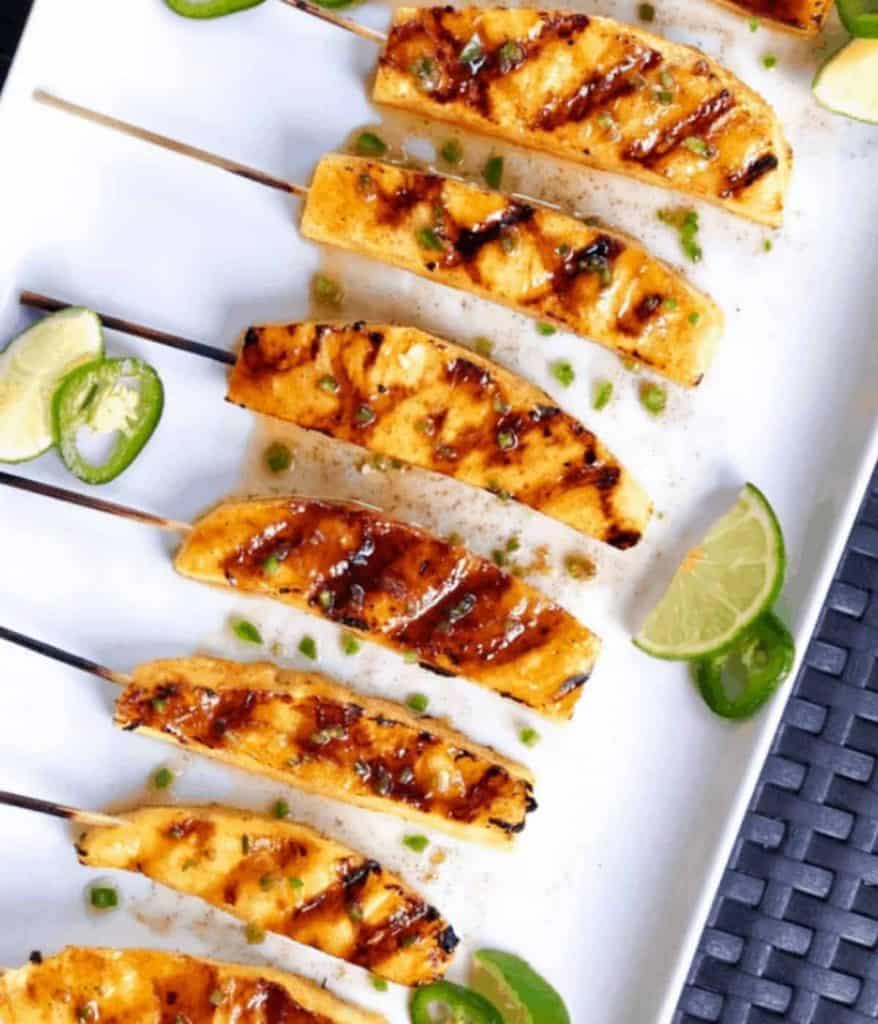A white plate with several skewers of grilled pineapple and some slime slices on the side.