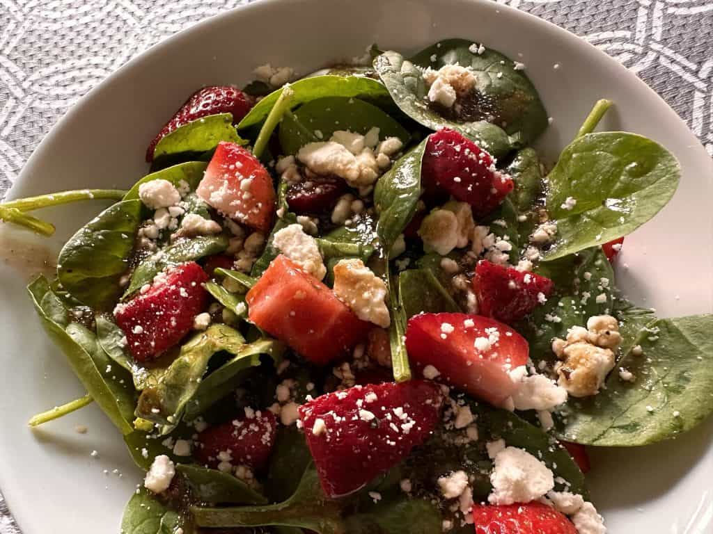 A white bowl with sliced fresh strawberries, baby spinach, and feta cheese topped with balsamic dressing.,