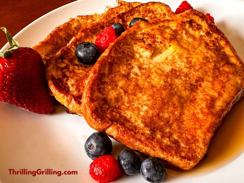 A white plate with three pieces of Blackstone French Toast covered in maple syrup and garnished with fresh berries.