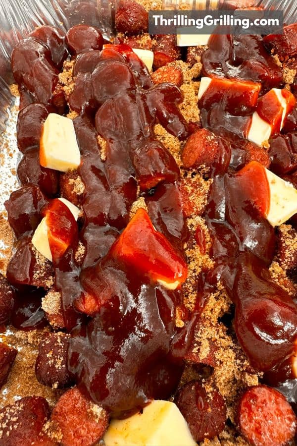 A tray of smoked hot dogs sliced into bite sized pieces with butter, brown sugar, and barbecue sauce on top.