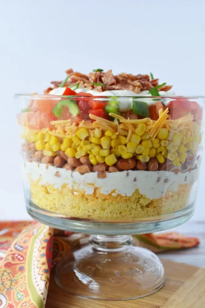 A trifle dish filled with layers of cornbread, cherry tomatoes, bacon, beans, and bacon