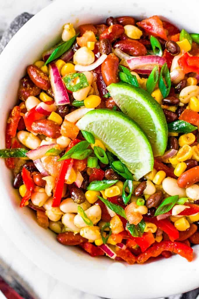 A white bowl full of 3 bean salad with beans, corn, onions, red peppers, and slices of lime.
