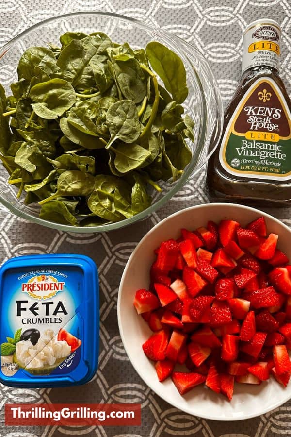 Salad ingredients laid out on a table :baby spinach, feta cheese, balsamic dressing, and fresh strawberries.