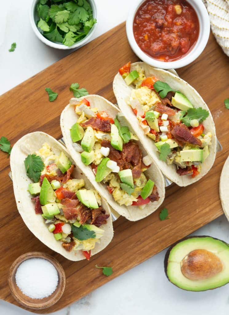 A cutting board with 3 breakfast tacos overflowing with eggs, bacon, avocado, and cliantro.