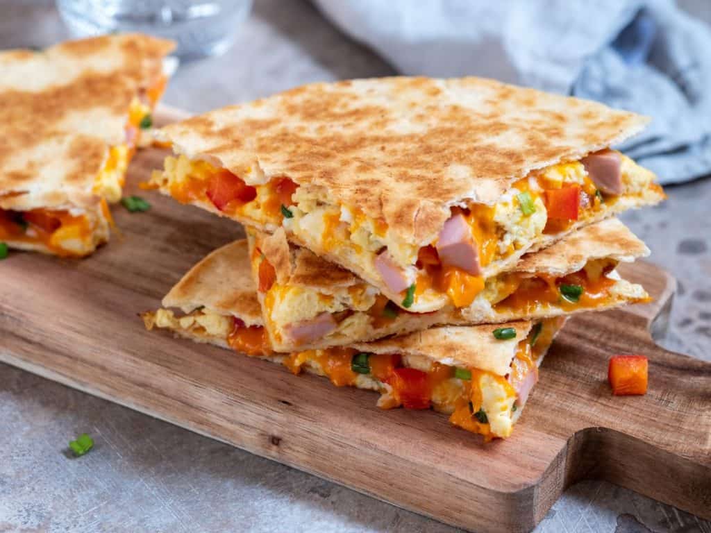 Small cutting board with 3 breakfast quesadillas stacked on top. They're bursting with scrambled eggs, cheese, ham, and red peppers and the tortilla is beautifully browned