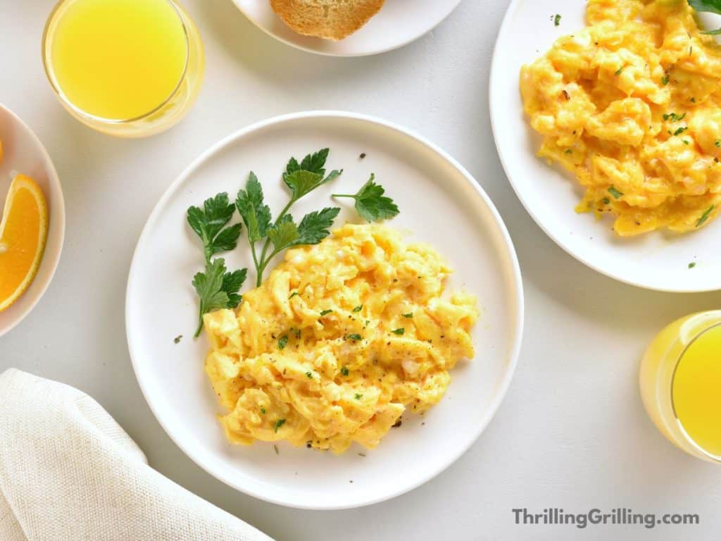 White plate with creamy scrambled eggs garnished with parsley. Also a glass of orange juice and an orange slice.