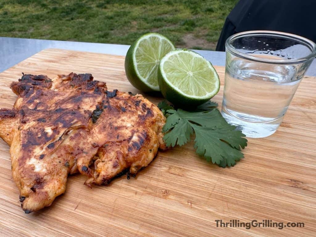 Wooden cutting board with an amazing piece of tequila lime chicken, two lime halves, a few cilantro leaves, and a shot glass of tequila.