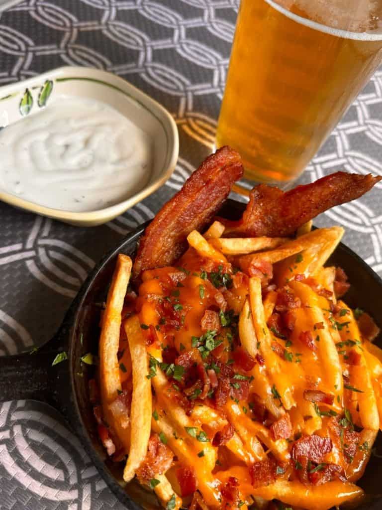 A small skillet loaded with bacon cheese fries, a small dish with ranch dressing for dipping, and a glass of beer. 