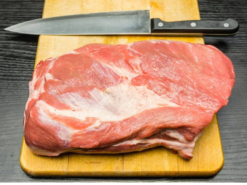 A pork butt sitting on a wooden cutting board with a sharp knife next to it.